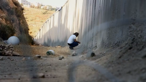 Still image from Israelism: a man squats down next to the West Bank Barrier.